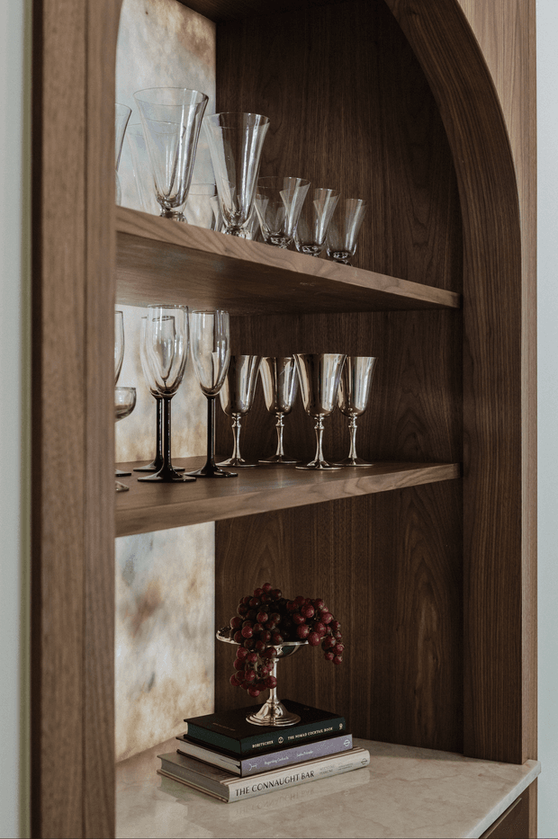 Close-up of a built-in arched display cabinet with dark timber interior and frame, two timber shelves, and a pale stone countertop. The shelves display clear and dark-stemmed wine and champagne glasses, while stacked books and a decorative bowl of grapes sit below. Warm, refined bar-storage styling.
