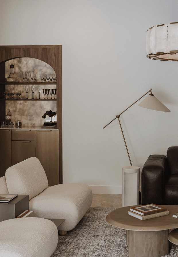 A refined living room vignette featuring a sculptural cream boucle lounge chair and ottoman, a dark leather sofa, a round wood coffee table, and a tall brass-toned floor lamp. A built-in dark timber bar cabinet with glassware and a stone-like textured backing occupies the left side, while a layered neutral rug and warm stone flooring create a sophisticated, softly lit setting. A partial sculptural pendant light is visible overhead.