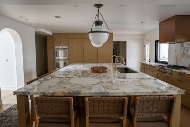 A spacious, refined kitchen centred around a large marble island with an integrated sink and brass-toned tapware. Warm timber cabinetry runs along the back and side walls, incorporating double ovens and additional storage. Three woven bar stools sit at the island, while a large globe pendant light hangs above. White walls, arched detailing, and dark-framed windows complete the bright, elegant interior.