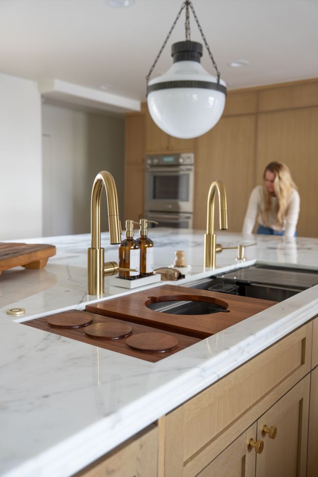 A refined contemporary kitchen centered on a large marble island with integrated dark sink, brushed brass gooseneck taps, timber cabinetry, and a dark timber sink-cover board with circular trivets. A globe pendant light hangs above, with built-in stainless-steel appliances and a person working in the background.