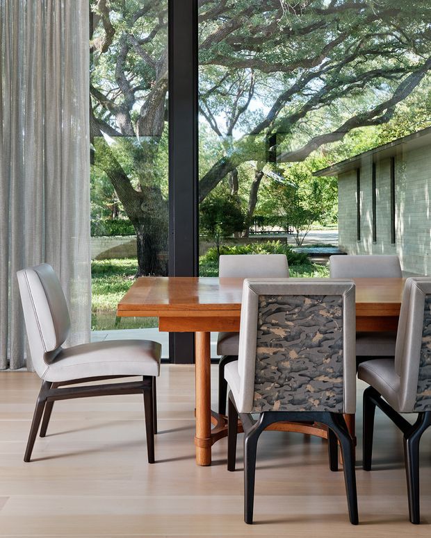 A refined dining room centered on a rectangular timber dining table and upholstered dining chairs. Full-height black-framed glazing opens views to mature leafy trees and a landscaped courtyard. Sheer curtains soften the glazing, while pale timber flooring and a light neutral palette create a bright, calm interior. A low, flat-roofed light brick or masonry structure is visible outside.