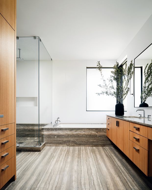 A bright, spacious contemporary bathroom featuring a freestanding bathtub clad in veined dark stone, a large frameless glass shower enclosure, double timber vanity cabinetry with a pale stone benchtop, and a full-height wall mirror. Vertical timber storage cabinetry and large white-framed windows add warmth and natural light. The floor and bath surround use strongly veined stone with grey, beige and brown tones.