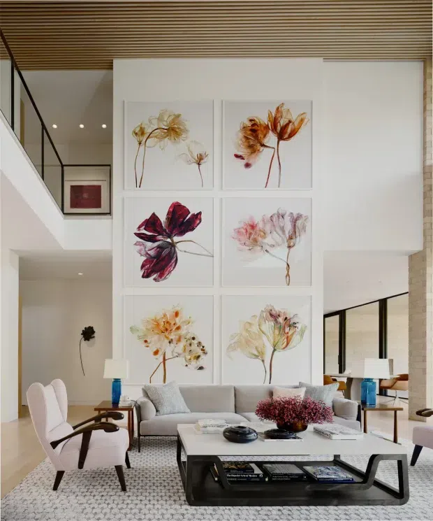 A bright, spacious contemporary living room with a large pale sectional sofa, sculptural armchairs, a substantial rectangular coffee table, and a patterned area rug. A six-panel botanical artwork arrangement forms the main feature wall. The room includes floor-to-ceiling glazing, a black-framed stair balustrade, an open mezzanine or upper-level landing, and a warm slatted timber ceiling. Neutral white and light-grey finishes are accented by blue table lamps and colourful floral artwork.