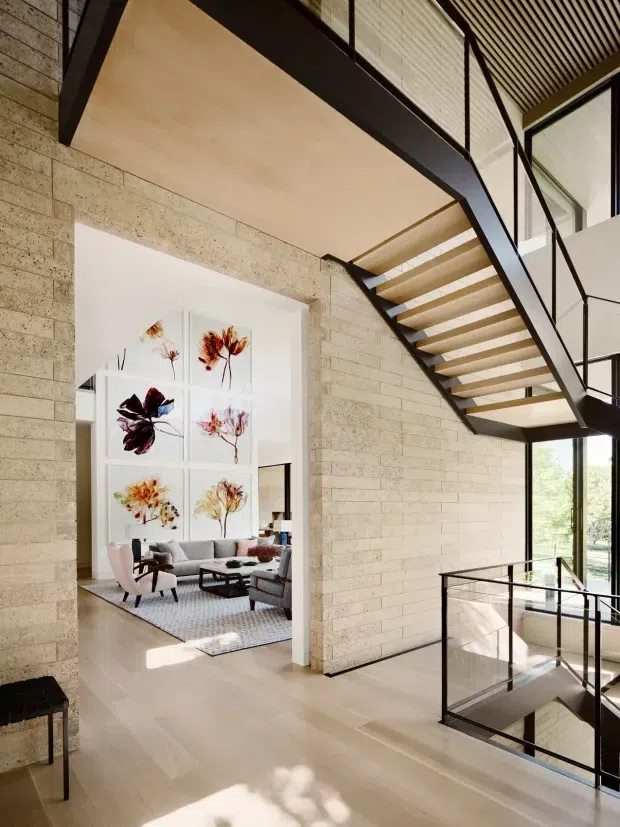 A bright, double-height interior landing or circulation space with a dramatic open-riser staircase, black metal structure and glass balustrades. Pale textured masonry walls, light timber flooring and a warm timber ceiling create a refined contemporary setting. A living room with sofas and framed botanical artwork is visible through the wide opening, while large windows bring in natural light.