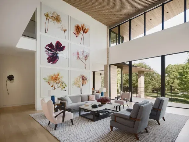 A spacious contemporary living room with a double-height glazed facade and mezzanine-like upper level. The room features a pale timber slatted ceiling, large black-framed windows, light timber flooring, a large patterned rug, a grey sofa and armchairs, a low dark coffee table, and a dramatic gallery wall of botanical artwork. The setting is bright, refined, and connected to the surrounding landscape.