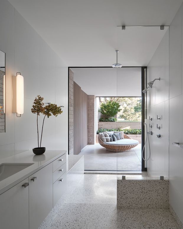 Bright contemporary bathroom with a walk-in shower and freestanding terrazzo bath in the foreground. White built-in vanity cabinetry runs along the left wall, while a large open glazed doorway connects directly to a landscaped outdoor terrace with built-in seating. The room features clean lines, pale surfaces, minimalist detailing, and abundant natural light.