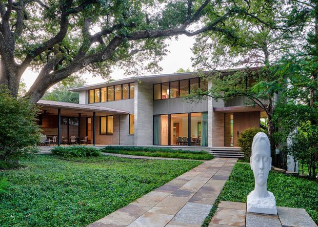 A modern two-storey residence set within a mature, heavily landscaped garden. The house features a rectilinear flat-roofed composition, extensive floor-to-ceiling glazing, warm timber wall panels, pale masonry walls, and sheltered terraces. A paved walkway leads through the garden toward the recessed entrance, with a prominent white sculptural head in the foreground.