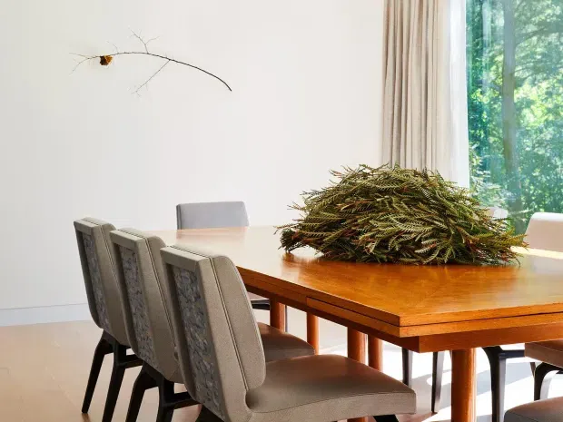 Bright dining room with a large polished timber dining table surrounded by upholstered dining chairs. Floor-to-ceiling windows with beige curtains bring in views of leafy greenery, while a minimalist branch wall sculpture and natural foliage centrepiece add organic accents.