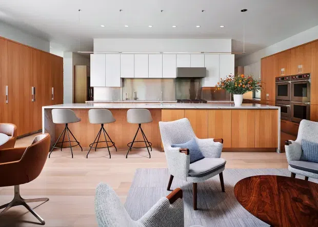 Open-plan kitchen and living area with extensive warm timber cabinetry, a long central island with seating, white upper cabinets, stainless-steel appliances and splashback, and mid-century-inspired lounge furniture around a dark wood coffee table. The space is bright, refined, and contemporary with strong timber detailing.