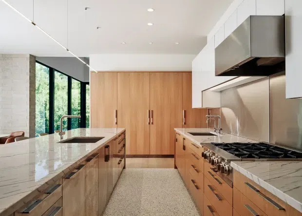 Contemporary, spacious kitchen with a large marble-look island, integrated sink and seating area, extensive timber cabinetry, stainless steel appliances, and a terrazzo-style floor. Full-height glazing along the side brings in abundant natural light and views of greenery.