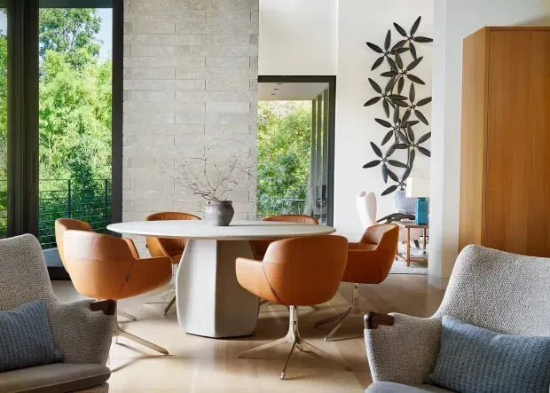 A bright, refined dining area with a large round white table and sculptural pedestal base surrounded by warm tan leather swivel chairs. Textured upholstered lounge chairs frame the foreground. The room features pale stone wall cladding, light flooring, expansive dark-framed glazing, a timber built-in door or cabinet, and a tall botanical metal wall sculpture. Lush greenery is visible outside through the windows.