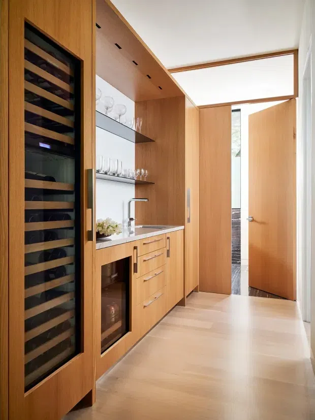 A compact, highly finished wine storage and serving area with full-height warm timber cabinetry, a glass-fronted wine refrigerator, open glassware shelves, a small sink, stone benchtop, and integrated drawers. The space opens toward a bright timber-lined hallway through several doors, with pale timber flooring and a clean contemporary fit-out.