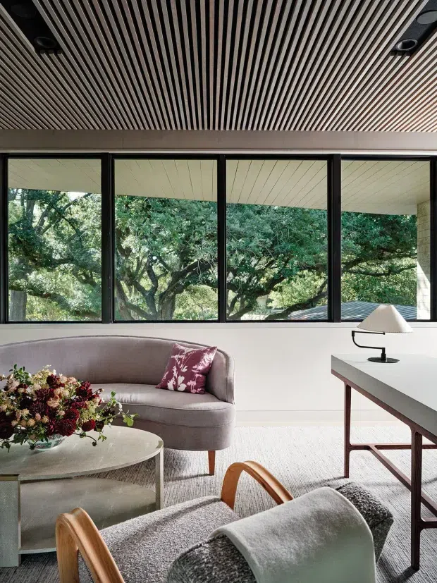 Contemporary living room with expansive black-framed glazing overlooking mature trees. The room features a curved taupe sofa, round coffee table, sculptural floral arrangement, a timber-framed lounge chair with draped throw, and a streamlined desk or console. A striking slatted timber ceiling with recessed lighting adds warmth and texture, while the restrained neutral palette creates a refined, calm atmosphere.