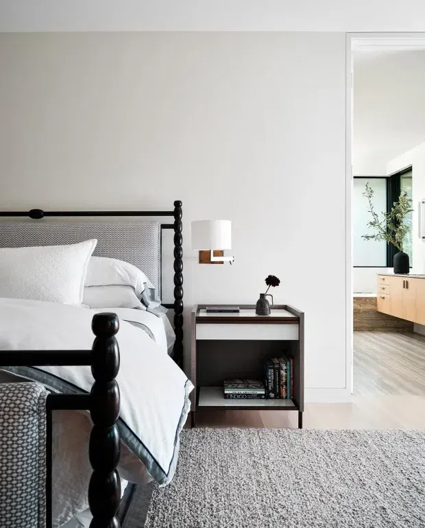 A refined bedroom with a dark turned-post bed, white bedding, a textured grey headboard and a compact dark timber bedside table. A wall-mounted modern lamp and neutral carpet create a calm monochromatic scheme. Through the open doorway, a timber-floored adjoining space with pale built-in cabinetry and dark-framed glazing is visible.