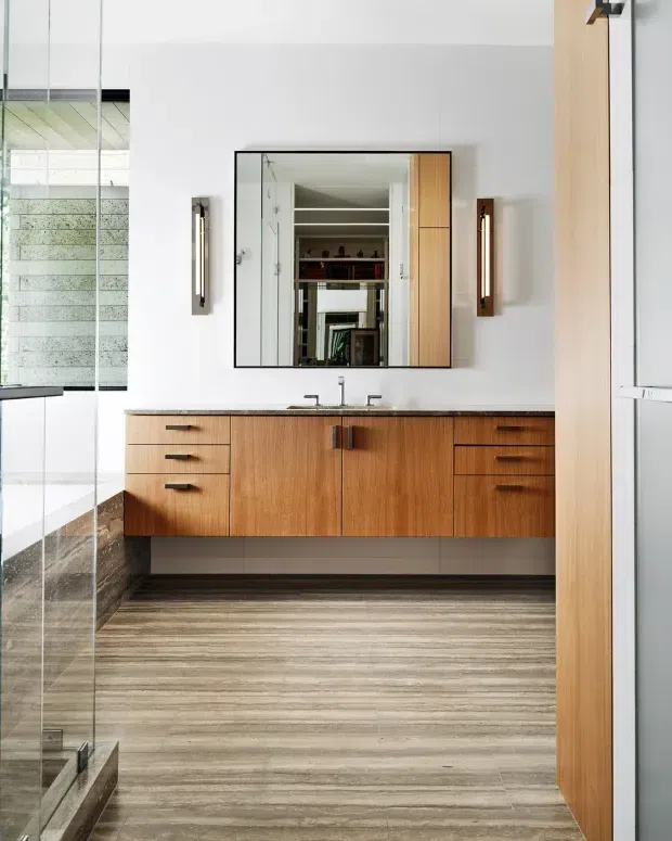 A contemporary bathroom vanity viewed through a frameless glass shower enclosure. The room features a wall-mounted warm timber vanity with drawers and double basins, a large rectangular mirrored cabinet, dark stone-look wall surfaces, and large grey-brown linear floor tiles. Reflections reveal additional shelving and bathroom details.