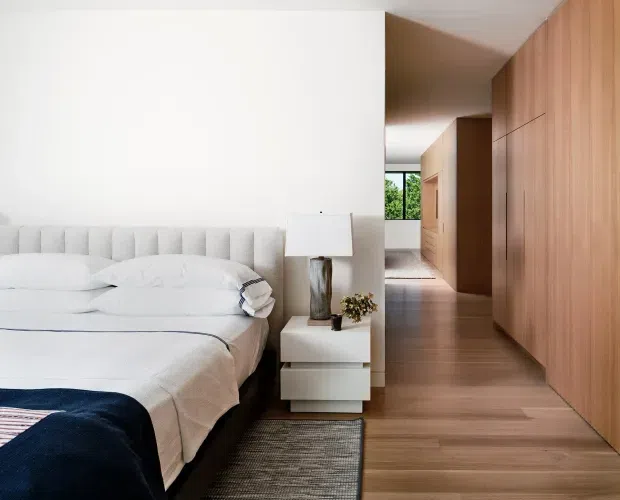 A contemporary bedroom with a low upholstered bed, white layered bedding, and a dark navy throw. A compact white bedside table with a sculptural lamp sits beside the bed. Warm timber flooring and extensive floor-to-ceiling timber built-in cabinetry line the corridor and right wall, creating a clean, integrated minimalist interior. A bright window at the rear provides greenery views.