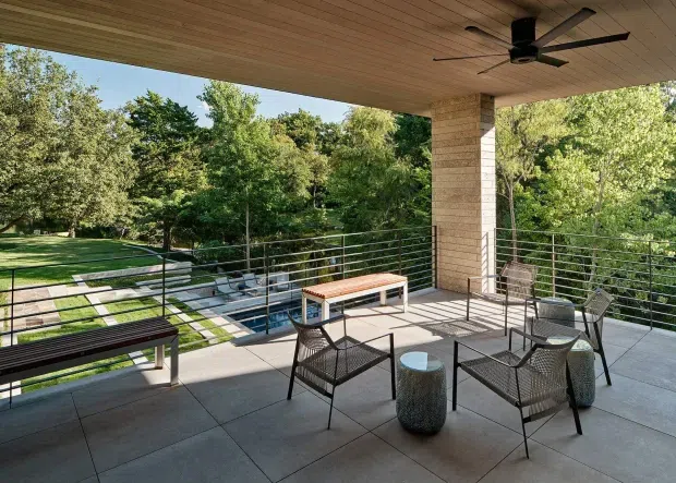 A spacious covered outdoor terrace overlooking a landscaped garden and swimming pool. The terrace features large-format grey paving, a timber-lined ceiling with a black ceiling fan, a pale masonry column, slim metal cable railings, and minimalist outdoor seating with benches, armchairs, and small side tables.