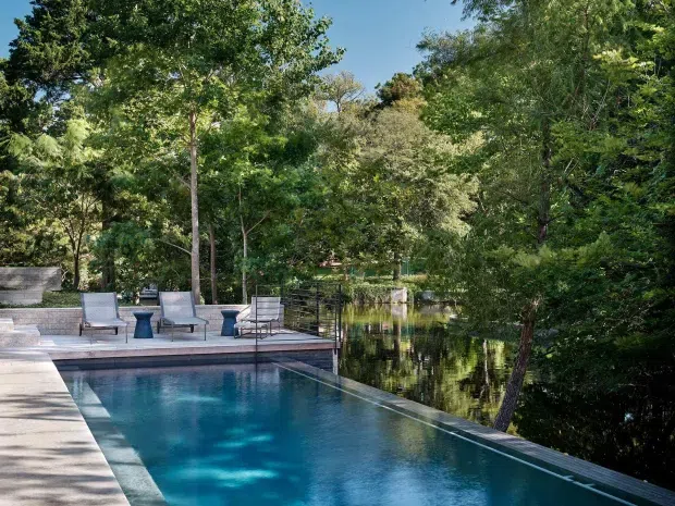 A landscaped outdoor pool area with a long rectangular swimming pool beside a calm wooded pond or waterway. The pool has pale stone or concrete surrounds, a timber-toned edge, and a small raised deck with contemporary outdoor lounge chairs and side tables. Dense mature greenery frames the scene, with strong reflections in the adjoining water.