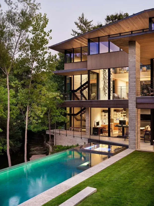 A multi-storey contemporary house set among mature trees, photographed from the landscaped pool terrace. The home features extensive floor-to-ceiling glazing, projecting flat roof planes, warm timber soffits, dark metal-framed windows and balustrades, vertical ribbed facade panels, and pale masonry columns. Interior living and dining areas are visible through the glazing, with a long narrow swimming pool and manicured lawn in the foreground.