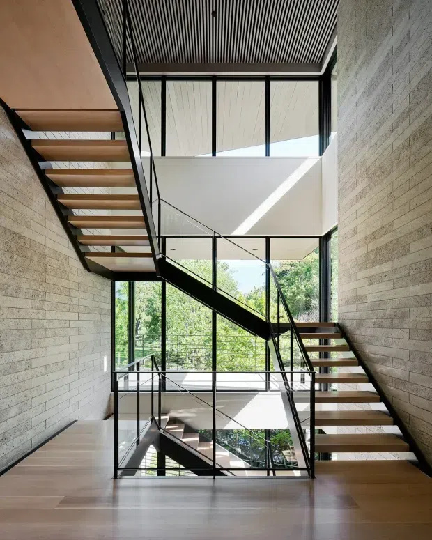 A bright, multi-level interior stairway with open-riser timber stairs, slim black metal framing and balustrades, and extensive glazing. Pale horizontal masonry walls contrast with warm timber flooring and stair treads. A dramatic open stair void and clerestory-like glazing bring in abundant daylight, with leafy trees visible outside.