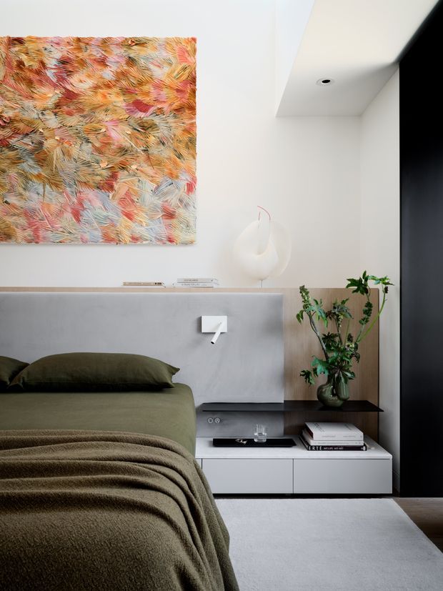 A refined contemporary bedroom with a low olive-green upholstered bed, layered textured bedding, and a floating white bedside cabinet. The room features a smooth grey half-height feature wall with integrated lighting, a large colourful abstract artwork, sculptural white vase, indoor plant, dark vertical joinery, and pale flooring. The palette is muted and sophisticated with organic green, grey, white, and warm multicoloured accents.