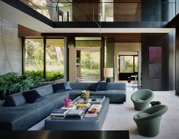 A sophisticated double-height contemporary living room with a large dark sectional sofa, sculptural green lounge chairs, and a substantial central coffee table. Full-height glazing opens the room to a leafy garden, while a mezzanine-level walkway with glass balustrades overlooks the space. The interior combines dark charcoal walls and joinery, warm timber ceiling panels, concrete surfaces, and abundant natural light.