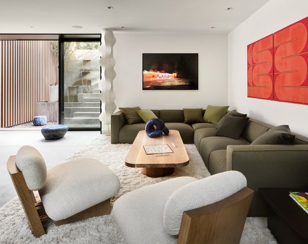 Contemporary living room with a low, deep olive sectional sofa, sculptural boucle lounge chairs, a rounded timber coffee table, pale textured rug, wall-mounted television, and bold red abstract artwork. Floor-to-ceiling glazing and vertical timber screening bring natural light into the space, with a sculptural open staircase visible at the rear.