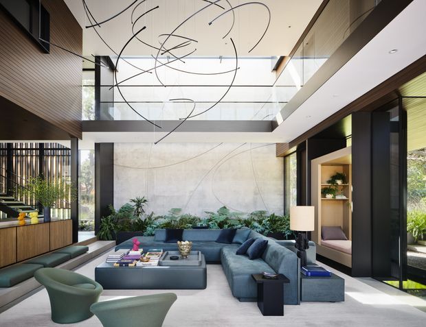 A refined contemporary living room with a double-height void and clerestory glazing. The space features a large blue-grey sectional sofa, sculptural green lounge chairs, a dark central coffee table, extensive indoor planting, timber wall and ceiling panelling, pale concrete-look surfaces, and expansive black-framed glazing. A suspended abstract linear light sculpture hangs overhead, while a planted courtyard or garden is visible through the glazing.