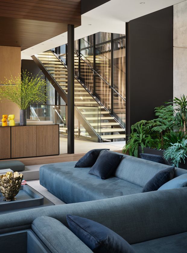 A sophisticated double-height living room with a large blue sectional sofa in the foreground, a dark coffee table, lush indoor plants, and a fluted timber console. A dramatic open staircase with glass balustrades and black framing rises through the space, backed by extensive floor-to-ceiling glazing. The interior combines dark feature walls, warm timber ceilings and joinery, concrete, and abundant natural light in a refined contemporary setting.