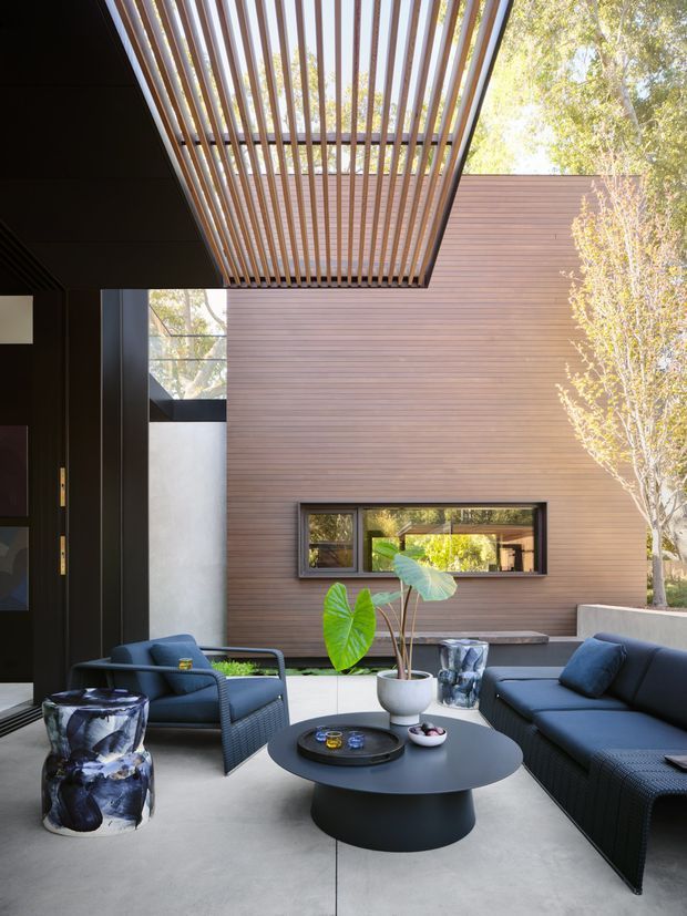 A contemporary covered outdoor lounge courtyard with dark woven sofas and armchairs arranged around a round black coffee table. The space features a smooth pale concrete floor, dark wall surfaces, a warm horizontal timber-clad wall with a narrow window, and a dramatic overhead timber slatted pergola. Potted tropical foliage and sculptural side tables add organic accents, while mature trees are visible beyond the courtyard.