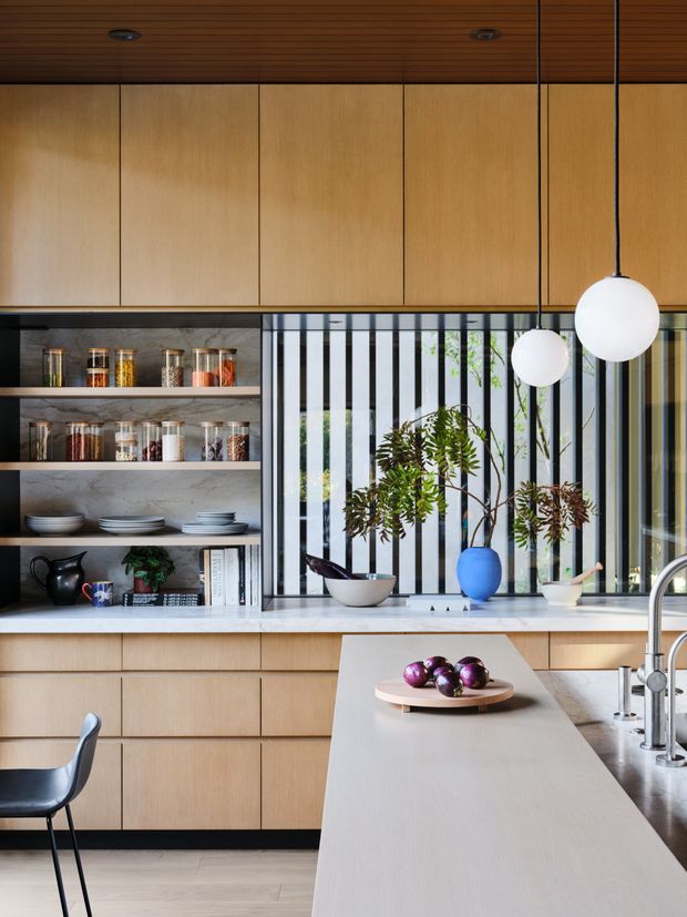 A refined contemporary kitchen with extensive light timber cabinetry, a timber slatted ceiling, open shelving, and a large pale island bench. The kitchen features a grey stone worktop and splashback, integrated stainless-steel sink and tapware, globe pendant lights, and a dramatic vertical black-and-white screened window behind the preparation area. Decorative plants, ceramics, and pantry jars add warmth and colour.
