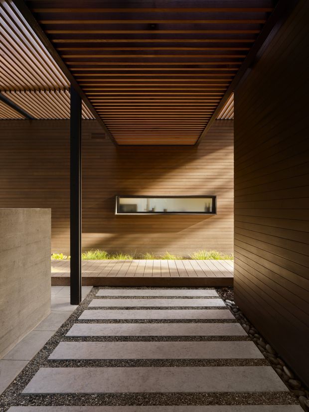 A sheltered architectural passage or entry courtyard with warm horizontal timber wall cladding and a slatted timber ceiling. Broad pale rectangular paving slabs are set into dark gravel, leading toward a raised timber deck and landscaped planting strip. A slim horizontal window, dark structural post, concrete wall, and carefully controlled natural light create a calm, highly crafted modernist composition.
