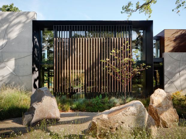 Front exterior entrance or boundary element of a contemporary property, framed by bold black rectangular walls and a tall timber vertical-slat screen. Large natural boulders, gravel, ornamental grasses, and a young tree create a landscaped approach, while glimpses of glazing and garden are visible behind the screen. Strong geometric composition with contrasting dark metal, warm timber, pale concrete, and natural stone.