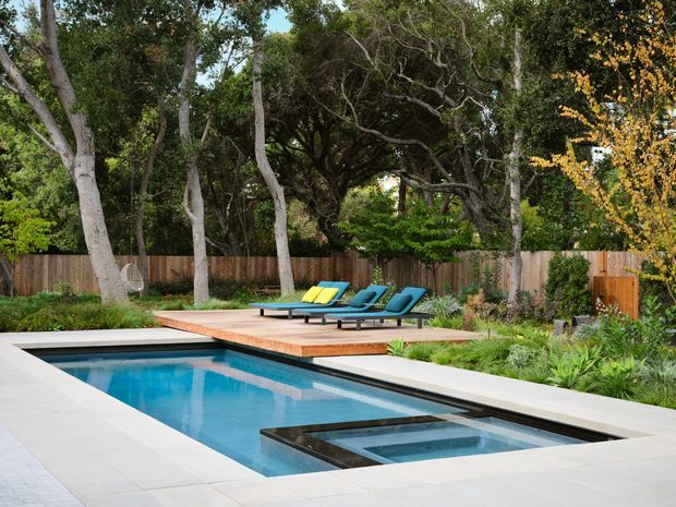 A landscaped backyard pool area with a rectangular swimming pool, integrated shallow spa or water feature, pale paved pool surround, and a raised timber deck with colorful lounge chairs. Mature trees, planted garden beds, and a timber privacy fence create a secluded outdoor setting.