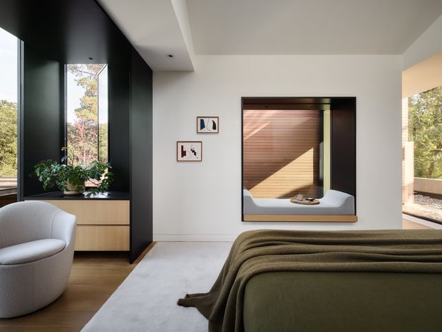 Contemporary bedroom with a low-profile bed dressed in an olive-green cover, a curved upholstered armchair, and light timber built-in cabinetry. A dark feature wall frames the window and cabinetry, while a recessed interior window reveals a warm timber-lined space beyond. The room has clean geometric detailing, abundant natural light, and views to surrounding greenery.