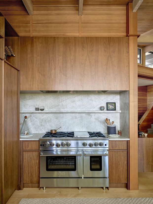 A compact, richly timber-lined kitchen focused on a large stainless-steel range cooker. Full-height warm wood cabinetry surrounds the cooker, with a pale grey stone backsplash and narrow display shelf. Exposed timber ceiling beams and wall panelling continue into the adjacent stair area, creating a crafted, warm interior with strong vertical and horizontal joinery lines.