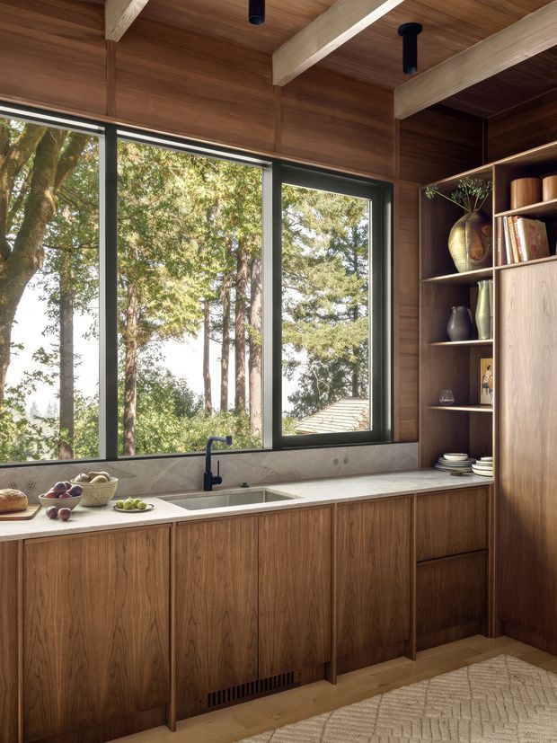 Warm, contemporary kitchen with extensive dark timber cabinetry and wall panelling, a pale stone benchtop, integrated sink and matte black tap. Large black-framed windows provide views into a wooded setting, while exposed pale timber ceiling beams and open shelving with ceramics add natural character.