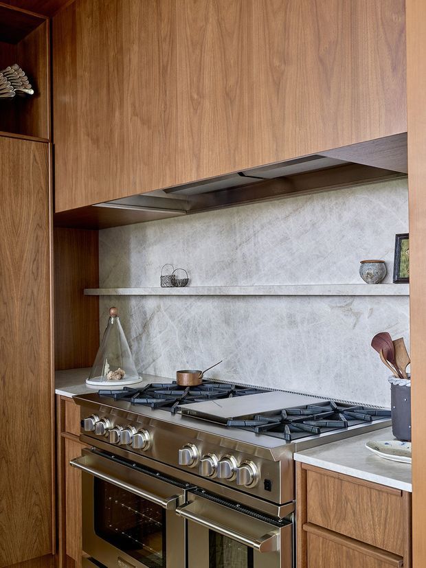 Close-up view of a sophisticated kitchen cooking zone featuring a large stainless-steel professional range with multiple gas burners and ovens, framed by extensive warm timber cabinetry. A pale veined stone backsplash and matching floating shelf provide a light contrast, with integrated stainless-steel extraction concealed beneath the upper cupboards. Small decorative objects and cooking utensils sit on the shelf and benchtops.
