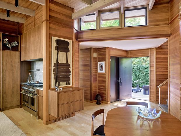 A warm, timber-lined open-plan interior combining a compact kitchen, dining area, and entryway. Exposed structural beams, extensive built-in timber cabinetry and wall panelling, clerestory windows, and a partially open mezzanine-like upper level create a richly crafted architectural character. The dining table occupies the foreground, while the kitchen and sculptural timber wall feature sit to the left and a glazed/open entrance leads outdoors at the rear.