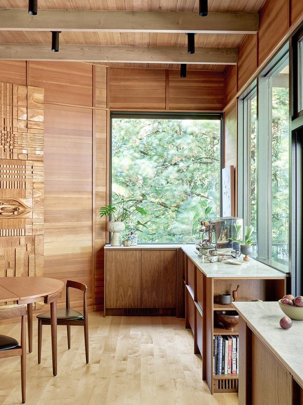 Warm, compact timber-lined kitchen and dining area with extensive natural wood wall and ceiling panelling, exposed structural beams, pale wood flooring, and large windows overlooking greenery. The kitchen has dark timber cabinetry, a pale stone benchtop, open shelving, integrated appliances, and a small round dining table with chairs. A sculptural textured timber feature wall adds visual interest.