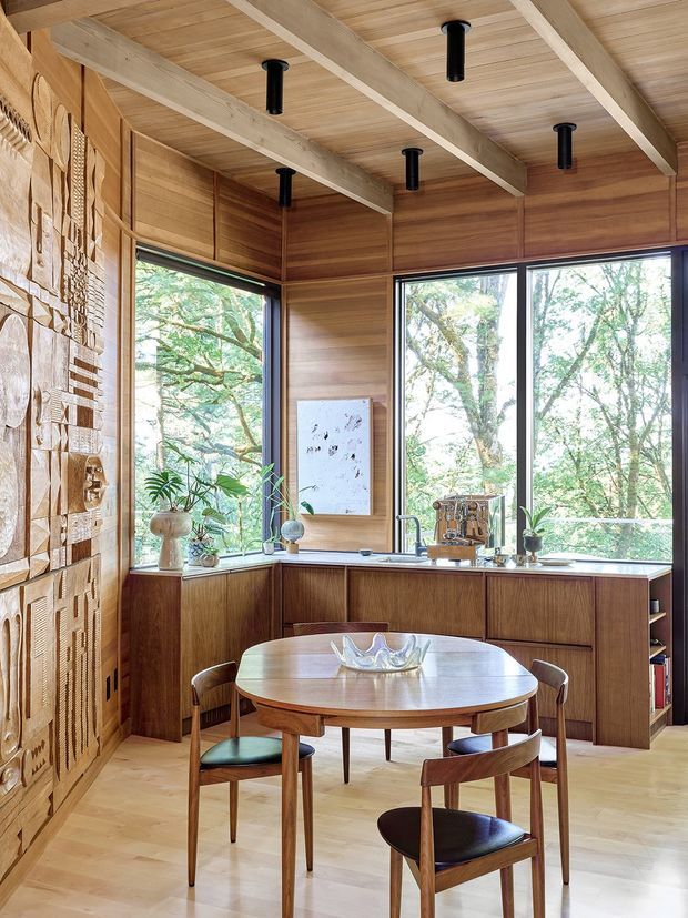 Warm, wood-lined dining area with a round timber table and mid-century-style chairs, set in front of a compact built-in kitchen or bar joinery. Floor-to-ceiling windows overlook leafy trees, while exposed pale timber ceiling beams, black cylindrical pendant lights, and a large textured carved wall feature add architectural character.