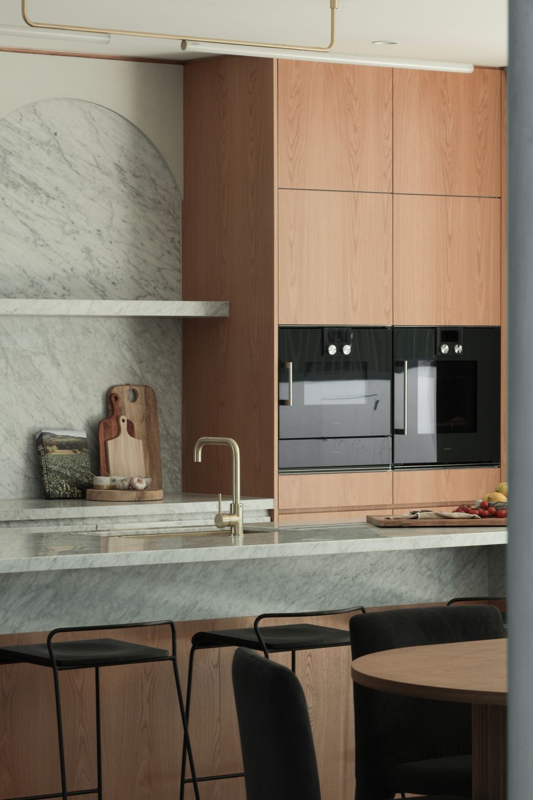 A central Alpine quartz stone wall anchors the design, complemented by a Carrara marble splashback and Gaggenau appliances. The adjoining wine bar framed by gentle curves creates a sense of refinement, balanced by travertine floors, white quartz pavers and marble enclaves. Details such as brushed brass tapware, green silk marble benchtops, and the Nood concrete pedestal bring character and intimacy, while textile lined robes and curved mirrors add a quiet elegance throughout.