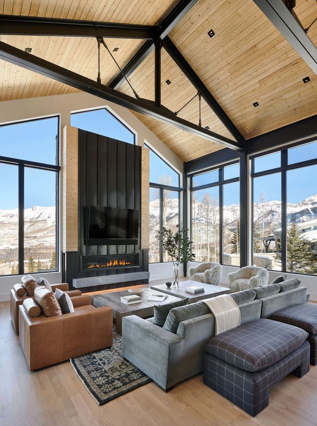 A spacious mountain-view living room with floor-to-ceiling glazing, a dramatic double-height vaulted timber ceiling, exposed dark structural beams, and a central black fireplace with television. The room is furnished with a large grey sectional sofa, brown leather armchairs, a coffee table, ottoman, patterned rug, and indoor plants. Snow-covered mountains are visible through the windows.