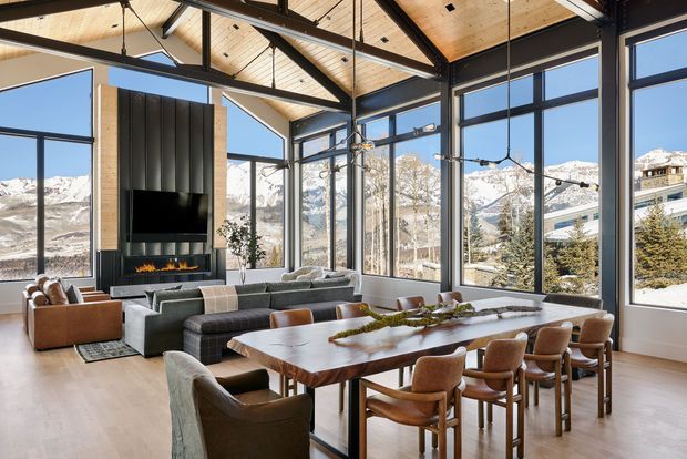 A spacious, double-height open-plan living and dining area with dramatic floor-to-ceiling glazing overlooking a snowy mountainous landscape. The room features exposed dark structural beams, a warm timber ceiling, a sculptural black fireplace wall with linear fire, a large timber dining table, upholstered dining chairs, and a generous sectional seating area. The overall design is refined, contemporary, and lodge-inspired.