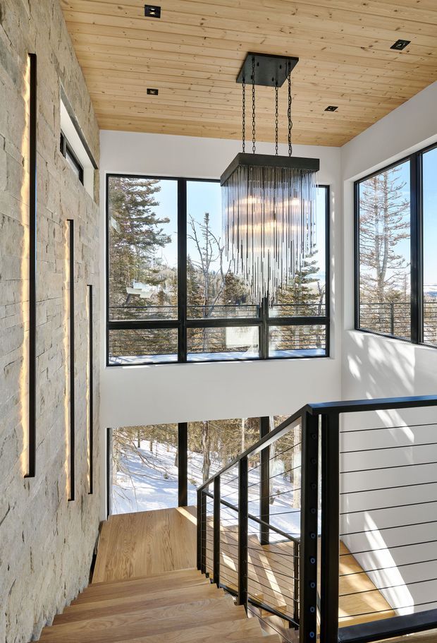 Bright contemporary stairway and landing with a tall glazed wall overlooking a snowy wooded landscape. The space features a warm timber-plank ceiling, pale textured stone wall, white walls, black-framed floor-to-ceiling windows, a suspended linear pendant with hanging metal rods, and a timber staircase with black metal and cable railing.