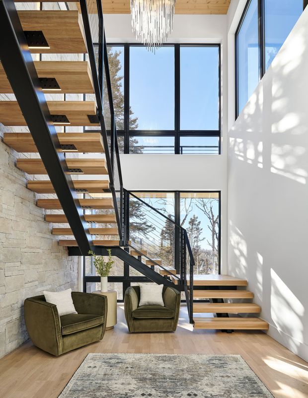 A bright, double-height interior stair hall with an open-riser floating staircase. The stair structure combines thick black steel stringers and balustrades with warm timber treads, set against a natural pale stone feature wall and white plaster walls. Large black-framed windows provide abundant daylight and views of trees, while a sculptural suspended chandelier hangs above. Two olive-green lounge chairs, a small side table, and a neutral patterned rug create a seating nook on the timber floor.