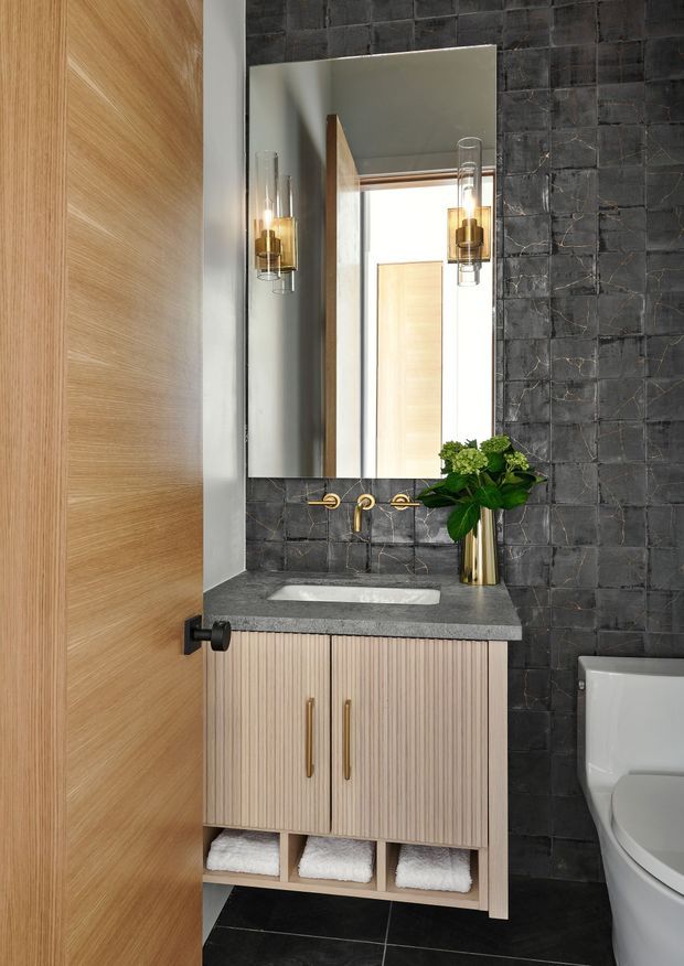 A refined compact bathroom vanity area viewed through an open timber door. The room features a floating fluted timber vanity with brass handles, a dark charcoal stone-look tiled wall, dark floor tiles, a grey stone benchtop with an integrated white basin, wall-mounted brass tapware, a large rectangular mirror, and matching glass-and-brass wall sconces. A white toilet is partially visible at right, with a green plant adding contrast.