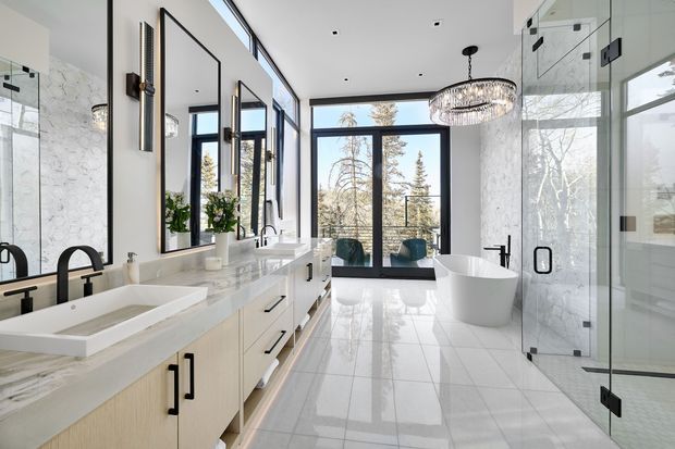 Bright, spacious contemporary bathroom with a long double vanity, pale wood cabinetry, white stone countertop, marble-look wall surfaces, black fixtures, a freestanding white bathtub, and a large glass shower enclosure. Floor-to-ceiling glazing and a glass door open the room toward a treed exterior, while a large crystal chandelier adds a refined focal point.