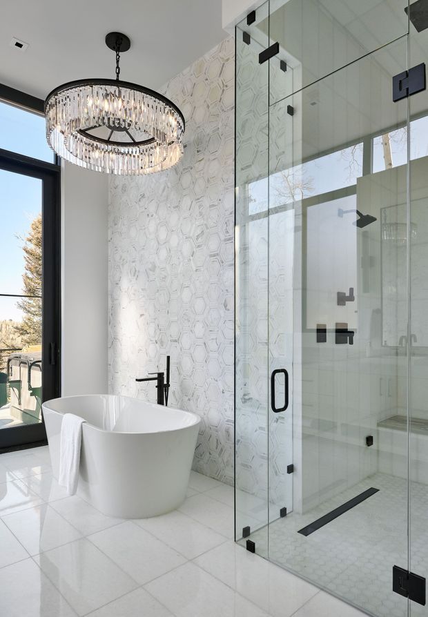 Bright, refined contemporary bathroom featuring a freestanding white bathtub beside a large frameless glass shower enclosure. The shower and feature wall use white geometric hexagonal-patterned tile, with pale large-format floor tiles and matte black fixtures and hardware. A dramatic circular crystal chandelier hangs from the ceiling, while a tall black-framed glazed door provides abundant natural light.