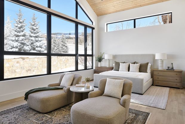 Bright contemporary bedroom with a large upholstered bed, two lounge chairs, an ottoman, bedside tables and layered rugs. Black-framed floor-to-ceiling corner glazing provides expansive views of snow-covered trees and a stone boundary wall. The room has white walls, a warm light timber ceiling, pale timber flooring and neutral beige furnishings.